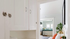 Une chambre avec un plancher en bois, des murs blancs, un lit avec des coussins orange et une commode blanche à tiroirs.