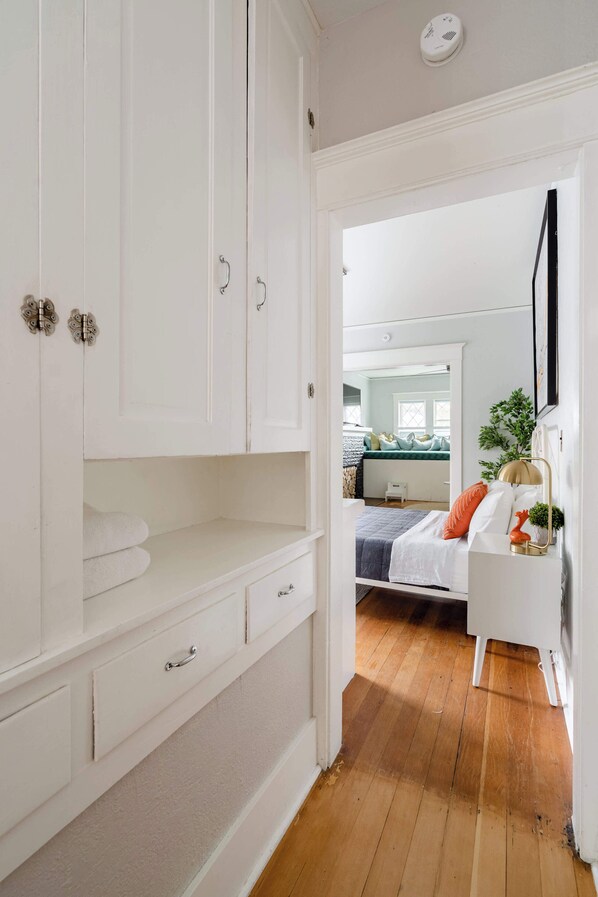 A bedroom with a wooden floor, white walls, a bed with orange pillows, and a white dresser with drawers.