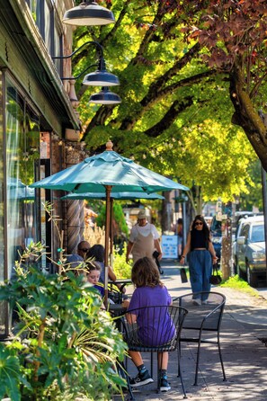 An outdoor cafe with tables and chairs under a green umbrella, a street with pedestrians, and parked cars.