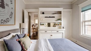 Un lit bien fait, recouvert d’une couvre-lit blanche et bleue, avec une tête de lit grise et des coussins décoratifs. Une commode blanche, dotée de tiroirs et d’étagères, se trouve à l’arrière-plan, et une fenêtre donnant sur l’extérieur est visible.