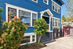 Una casa azul con detalles blancos, un porche y un pequeño árbol.