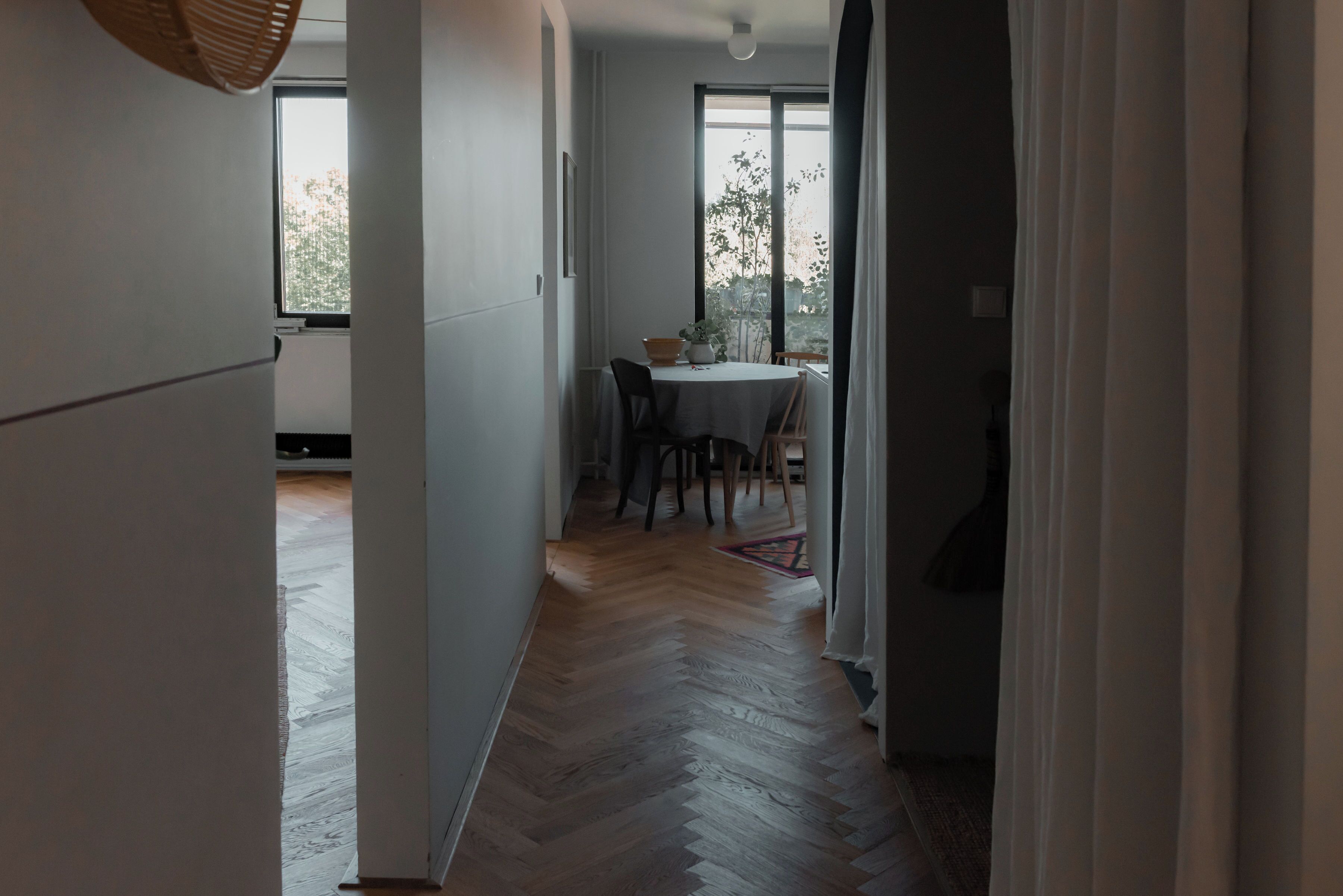 A hallway leading to a dining area with a table and chairs, wooden flooring, and a window with a view of trees.
