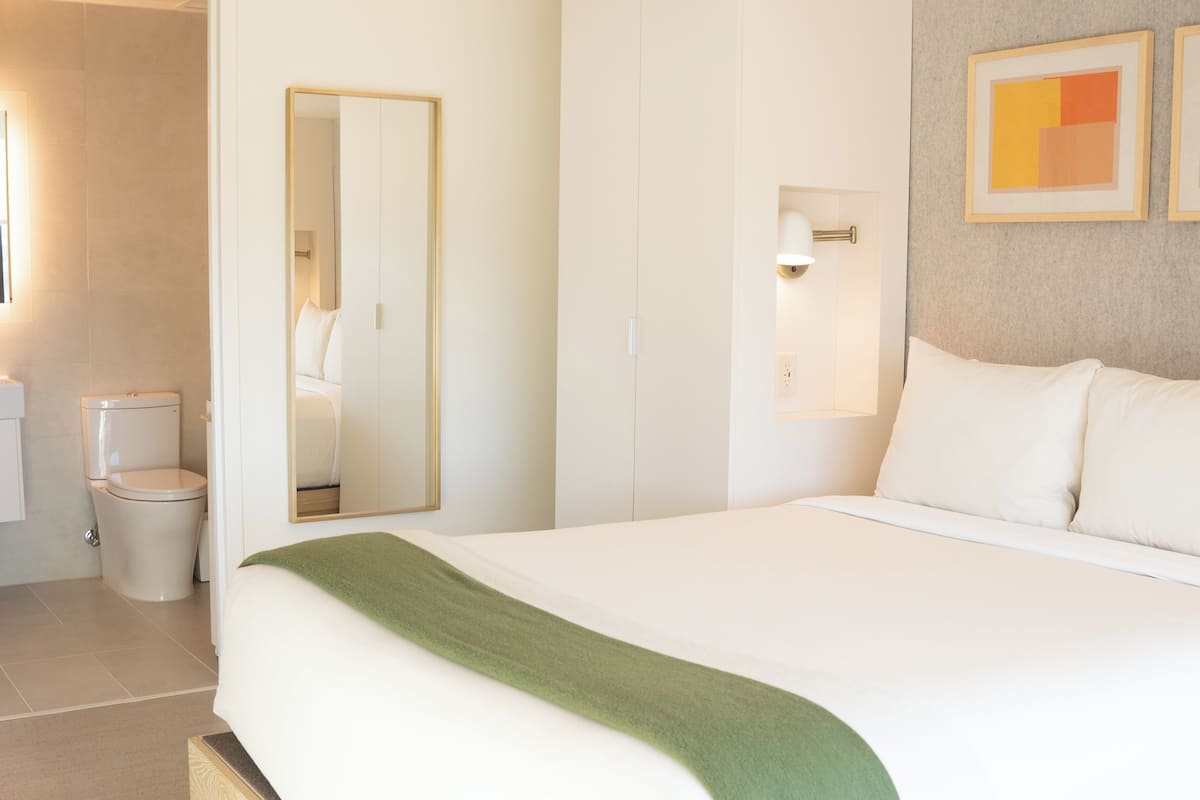 A hotel room with a bed, a mirror, a toilet, and a wall-mounted light fixture.