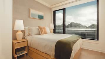 A modern bedroom with a large bed, two bedside tables with lamps, a bookshelf, a painting on the wall, and a view of a bridge through a large window.