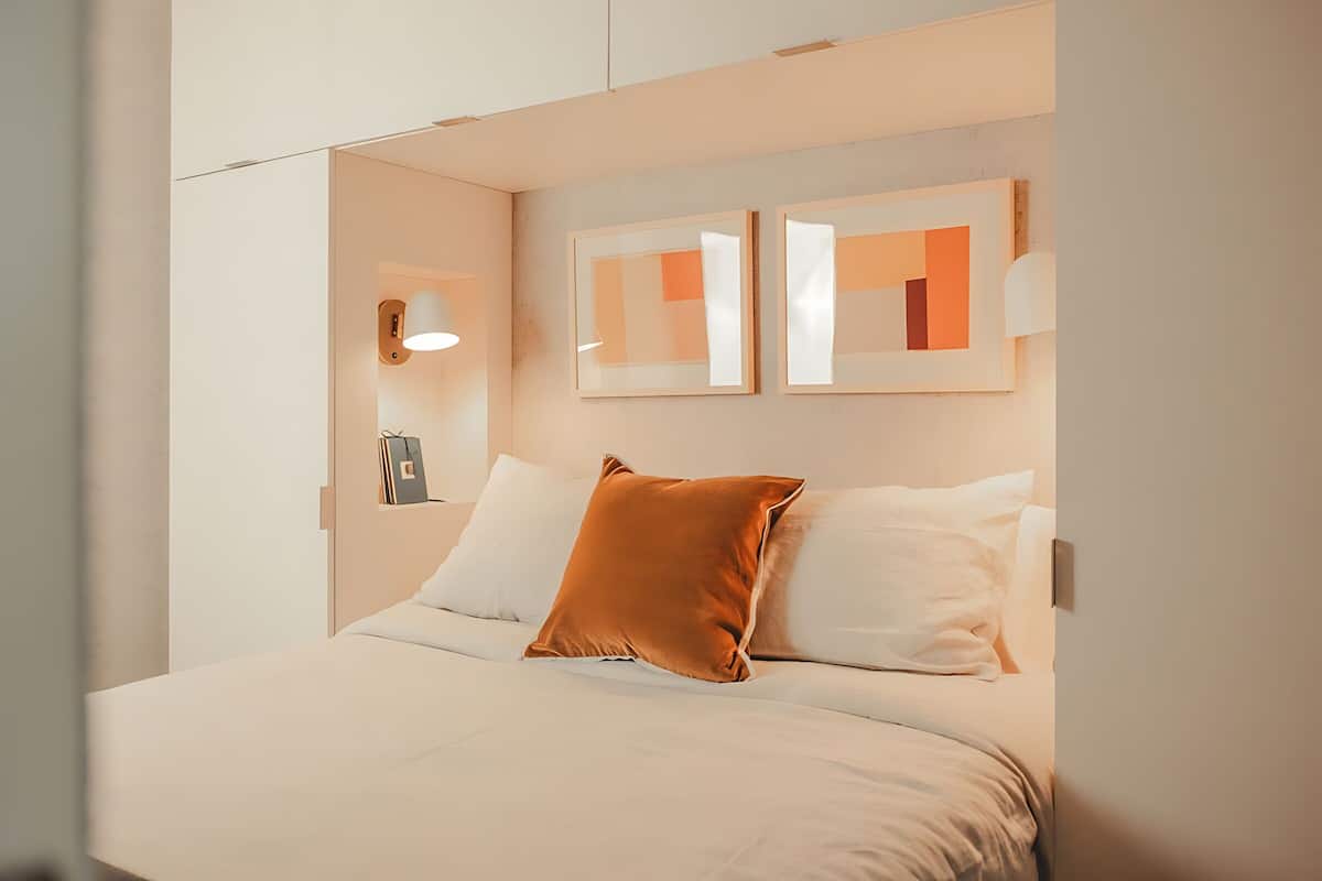 A modern bedroom with a large bed, white bedding, and a single orange pillow. There are two framed abstract paintings on the wall above the bed.