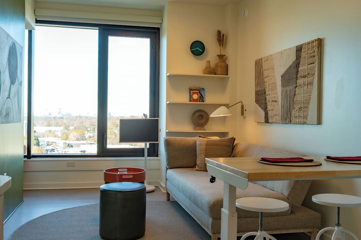 A modern living room with a large window, a sofa, a small table, and a round ottoman.
