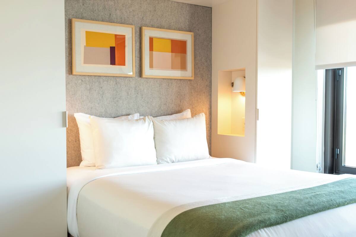 A hotel room with a large bed, two framed artworks on the wall, a bedside lamp, and a window with a view.