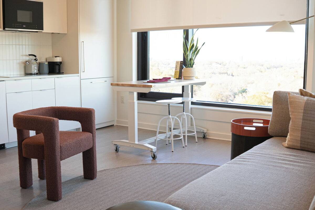 A modern kitchen with a microwave, kettle, and coffee maker. There is a window with a view of the city and a plant on the windowsill.