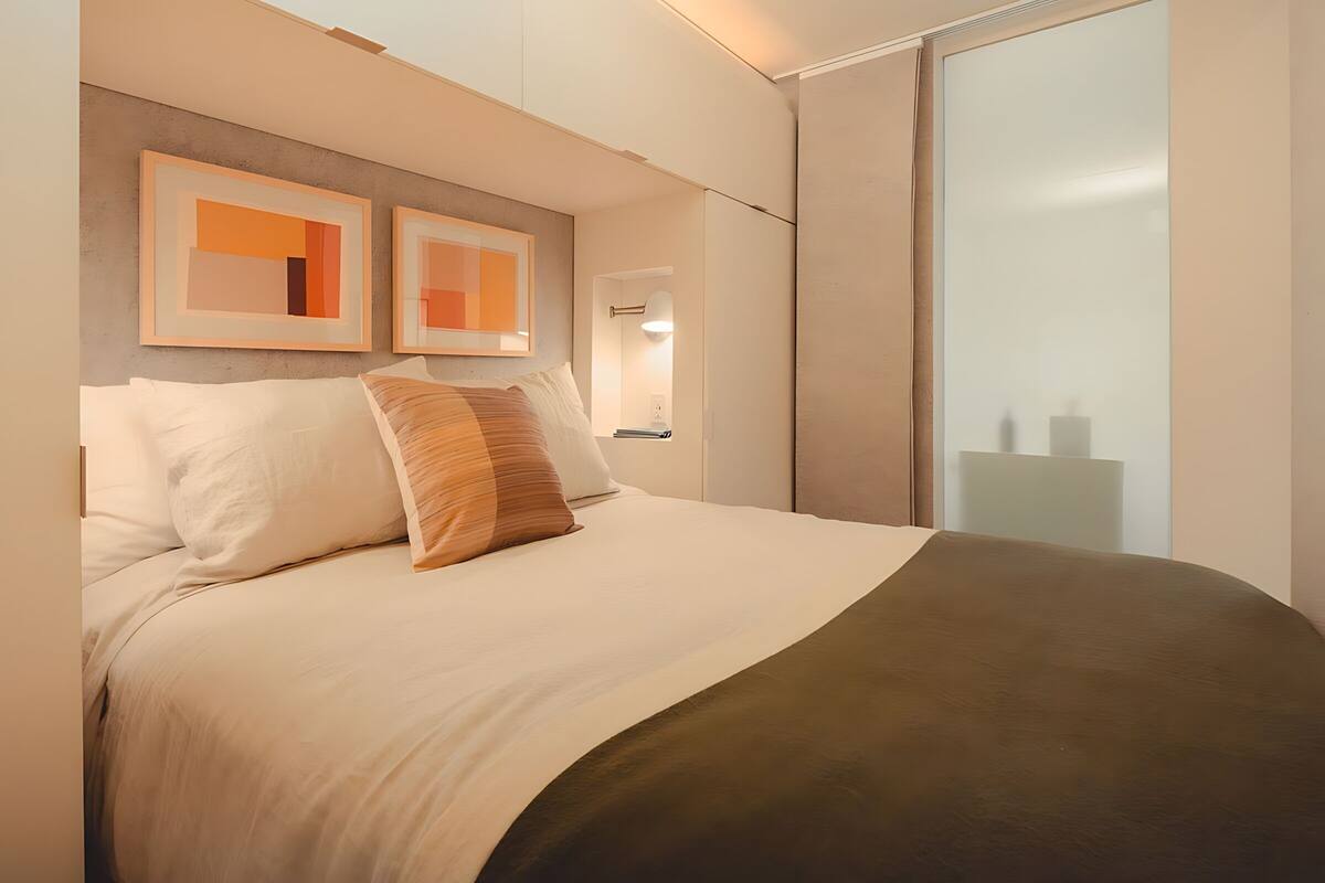 A modern hotel room with a large bed, two framed artworks above the headboard, a bedside table with a lamp, and a sliding glass door leading to a balcony.