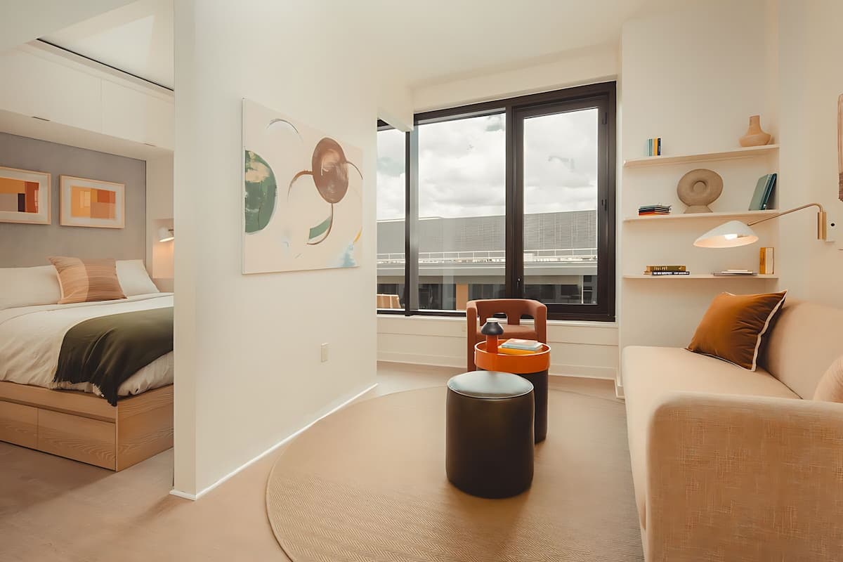 A modern hotel room with a bed, a sofa, a round rug, and a window with a view of a building.