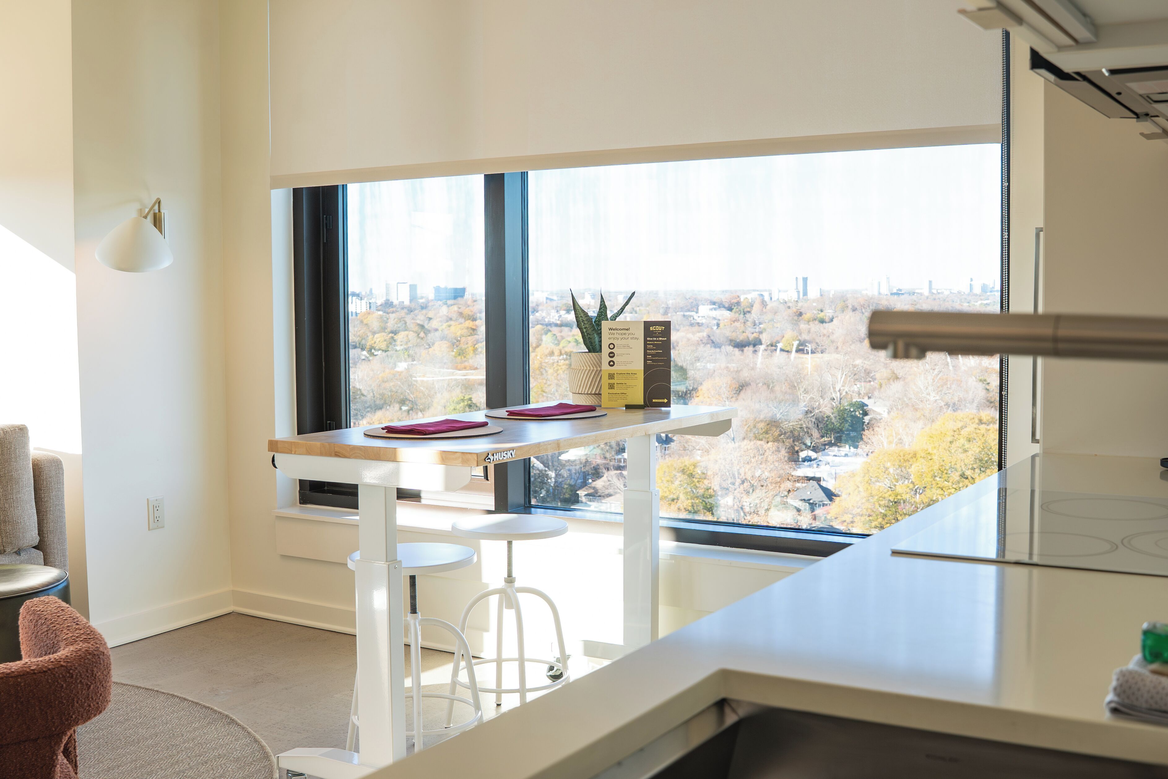 A modern kitchen with a large window, a bar stool, a small table, and a potted plant.
