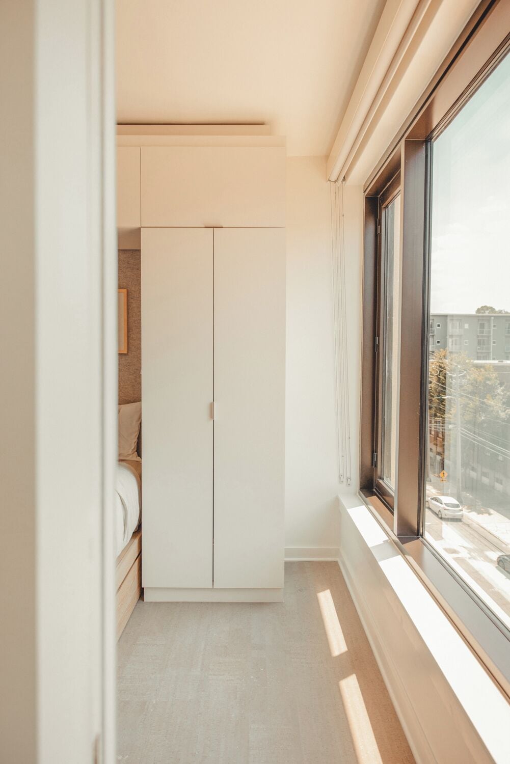 A narrow hallway with a white wardrobe, a bed, and a large window with a view of buildings and a car outside.