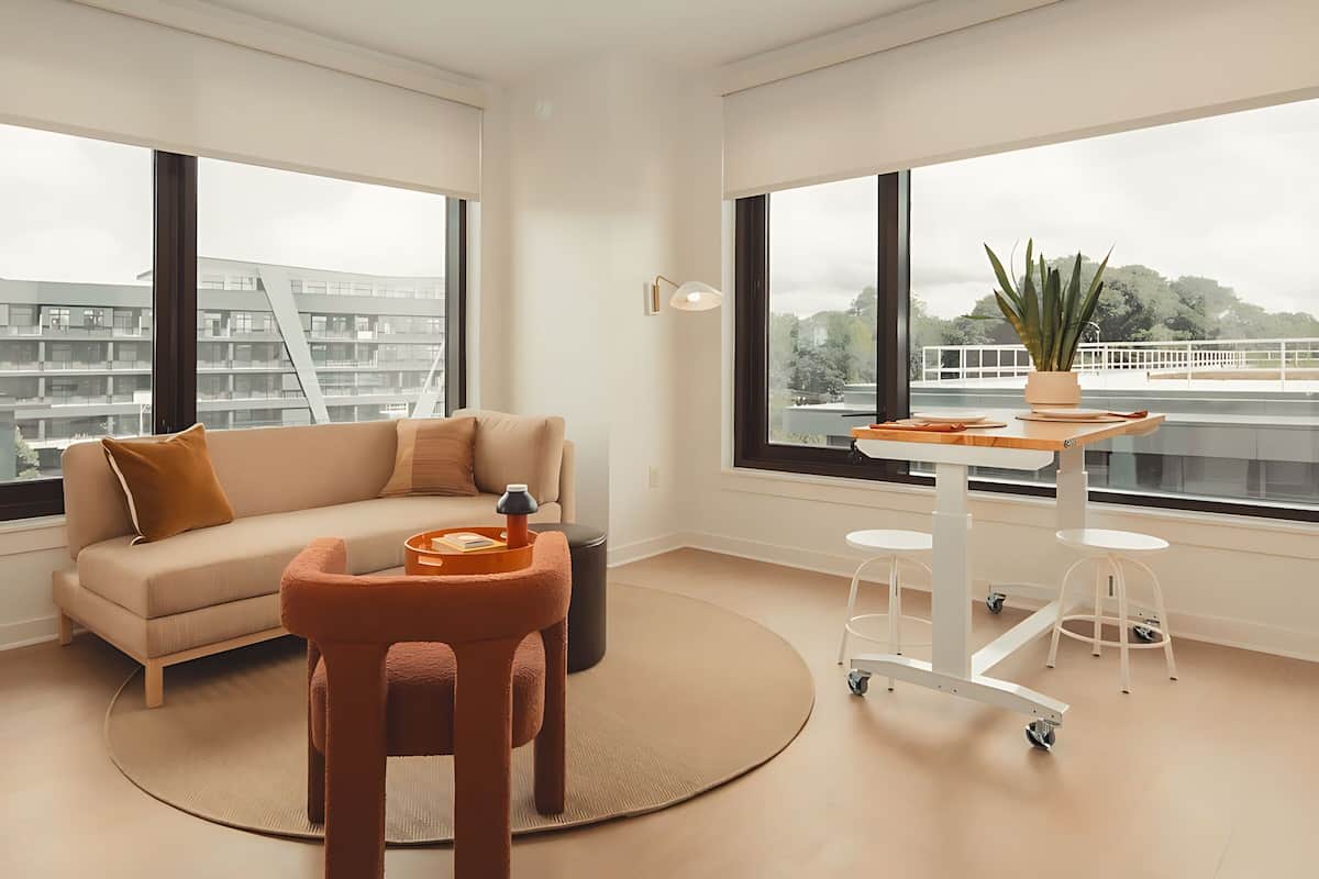 A modern living room with a sofa, a small round table, and a coffee table. There are large windows offering a view of a cityscape.