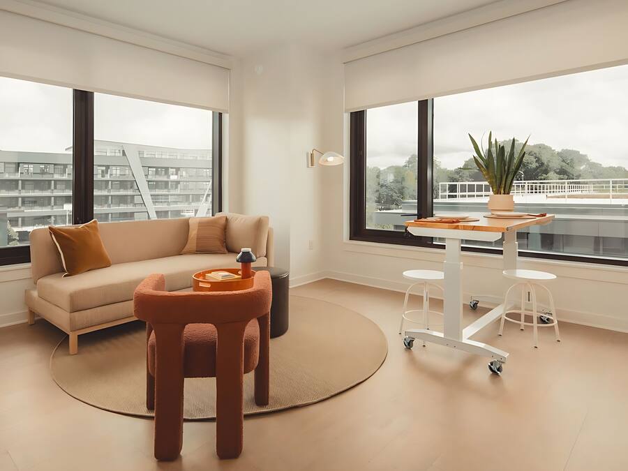 Una sala de estar moderna con un sofá, una mesita redonda y una mesa de centro. Amplias ventanas ofrecen vistas a un paisaje urbano.