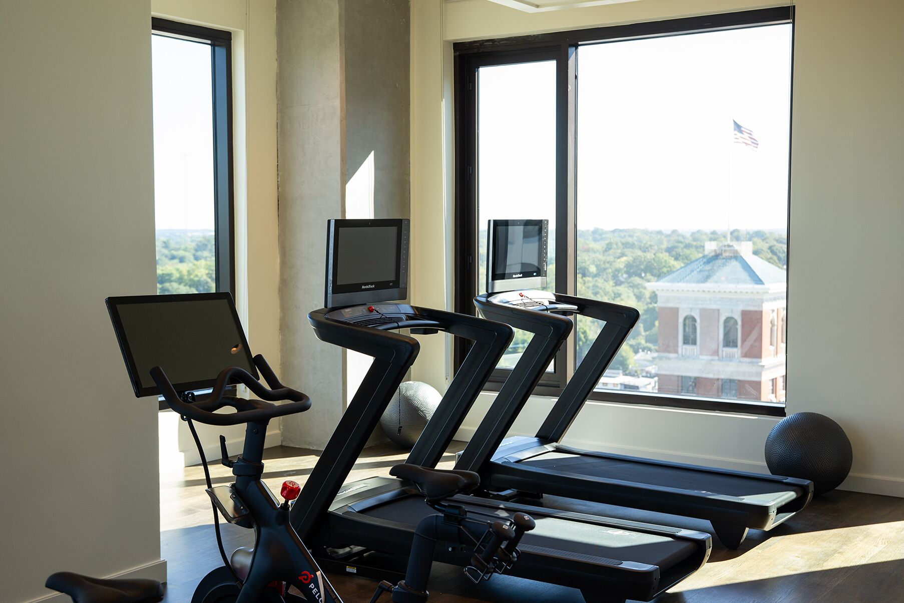 A home gym with treadmills and a stationary bike, large windows with a view of a building and trees.