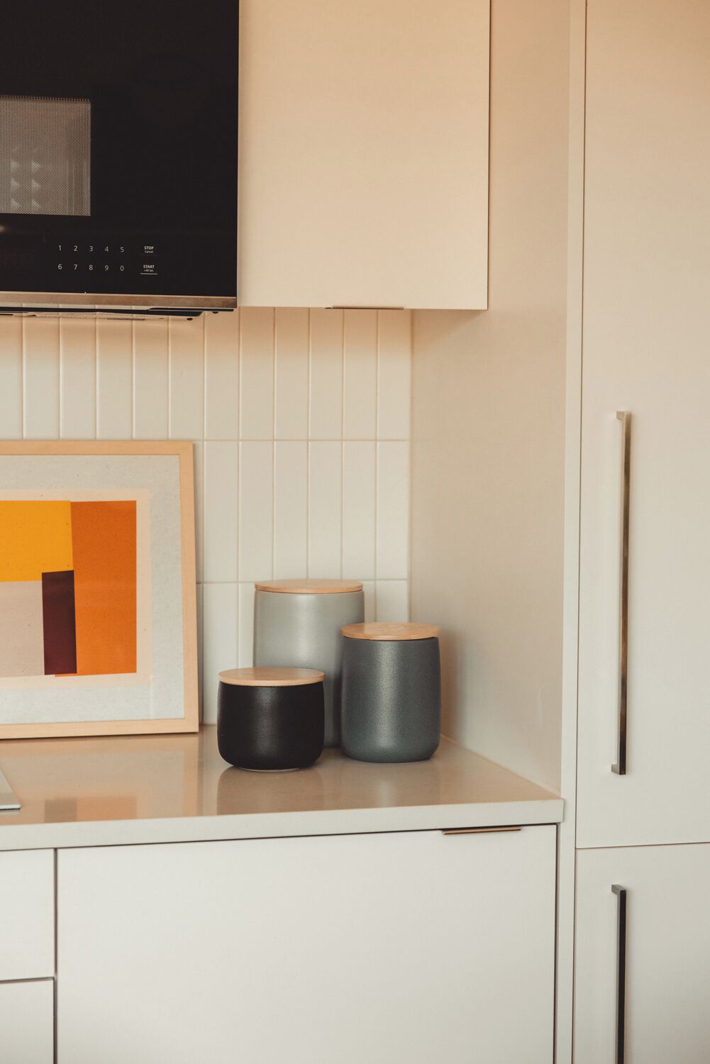A kitchen with white cabinets, a framed abstract painting, and three cylindrical containers on the counter.
