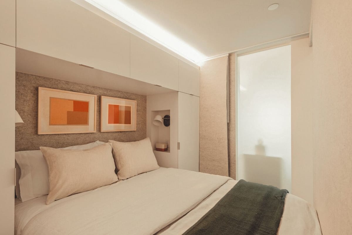 A modern bedroom with a large bed, two framed artworks on the wall, a bedside table with a lamp, and a sliding glass door to the right.