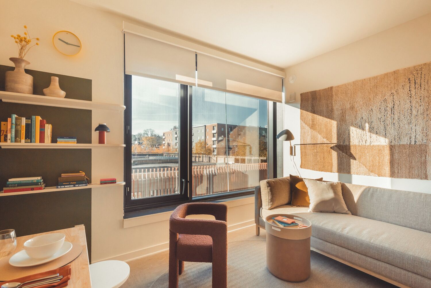A modern living room with a sofa, a coffee table, a bookshelf, and a large window offering a view of buildings.
