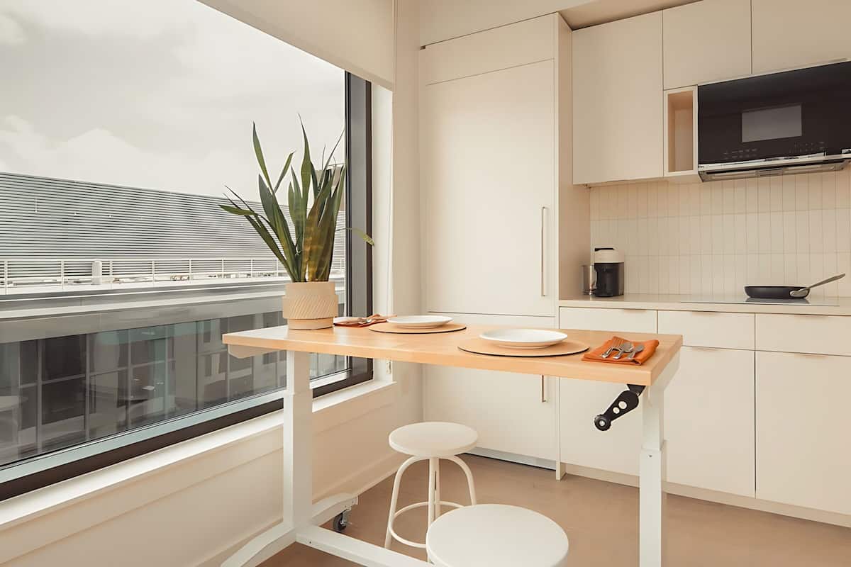 A modern kitchen with a wooden table, white stools, a potted plant, and a view of the city through large windows.