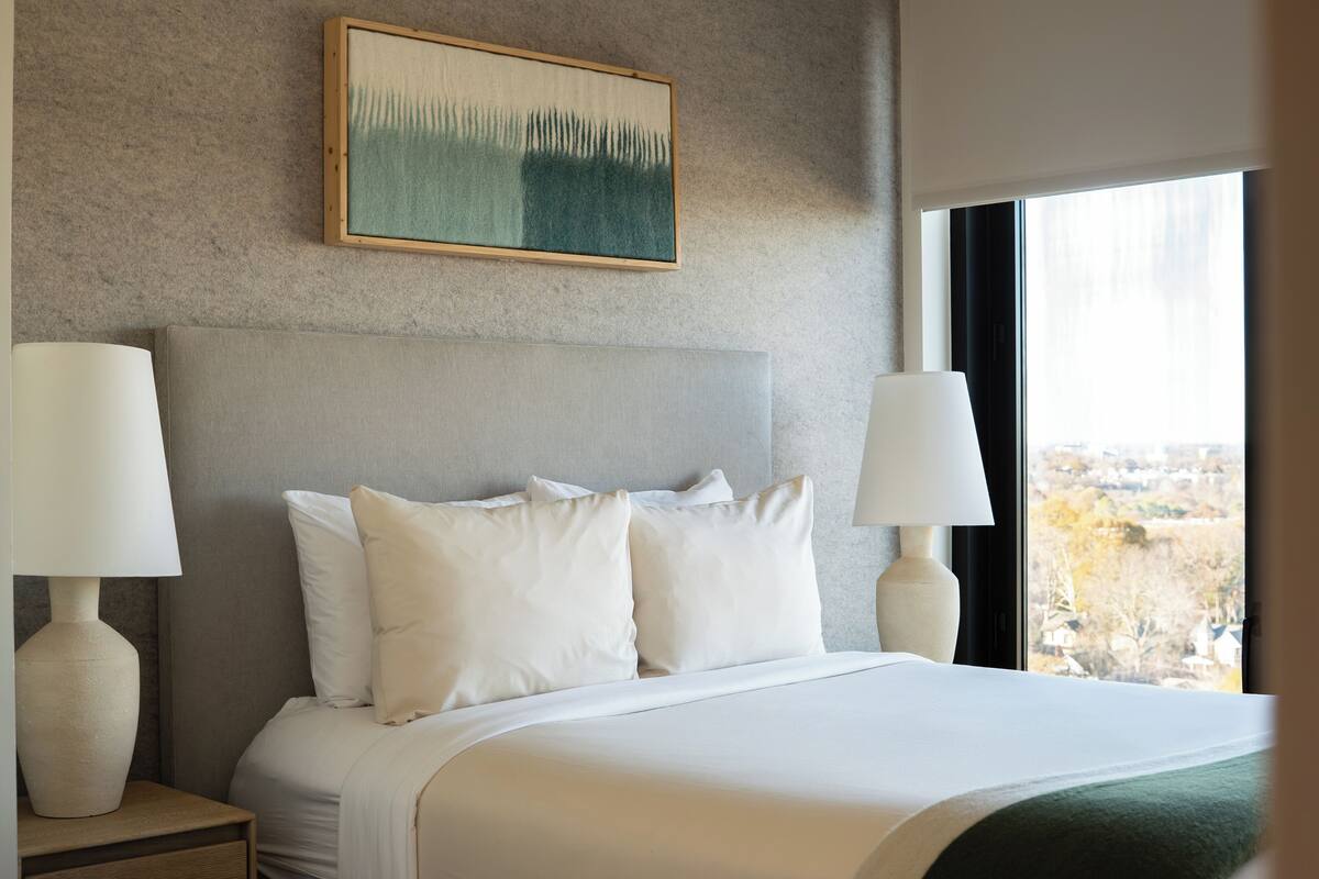 A hotel room with a large bed, two bedside lamps, a framed artwork above the bed, and a window with a view of a cityscape.