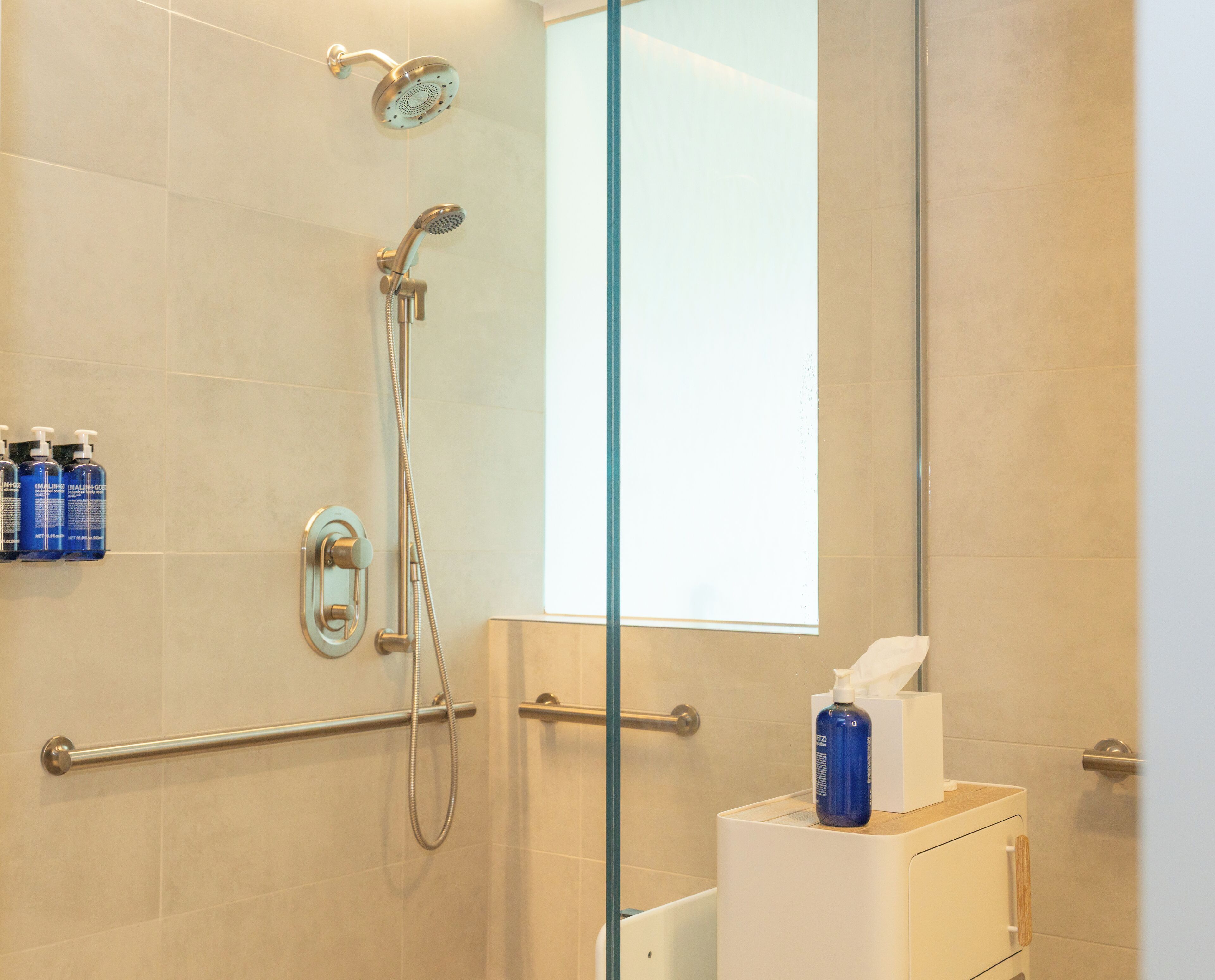 A modern bathroom with a glass shower enclosure, a handheld showerhead, a wall-mounted faucet, and a toilet paper dispenser.