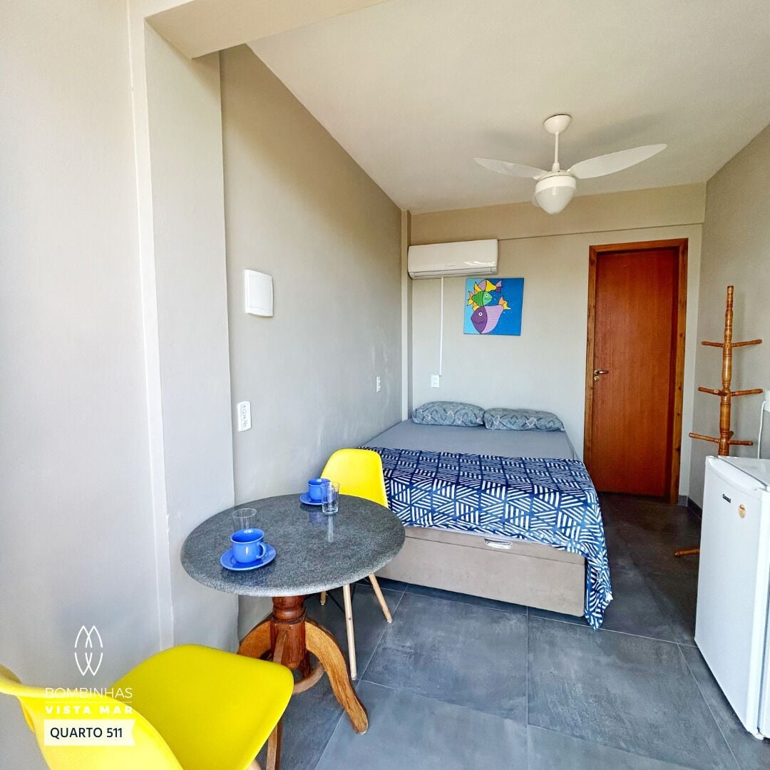 A compact bedroom with a bed, a small table with a cup, a chair, a ceiling fan, and a door.
