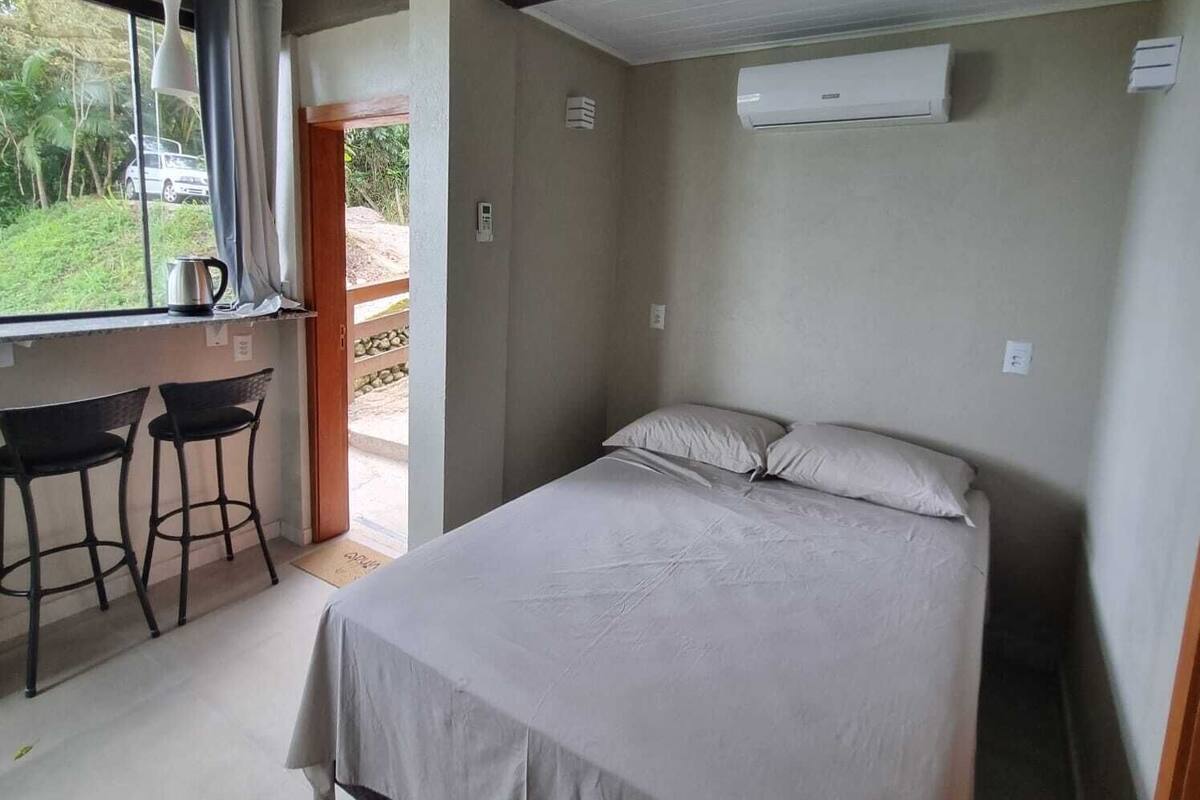 A bedroom with a bed, a desk with two chairs, a window with curtains, and an air conditioning unit.