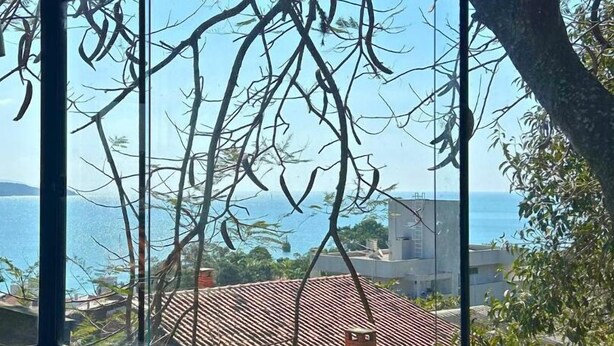 A room with a large window offering a view of the sea and buildings. - Bombinhas Vista Mar (Bombinhas)