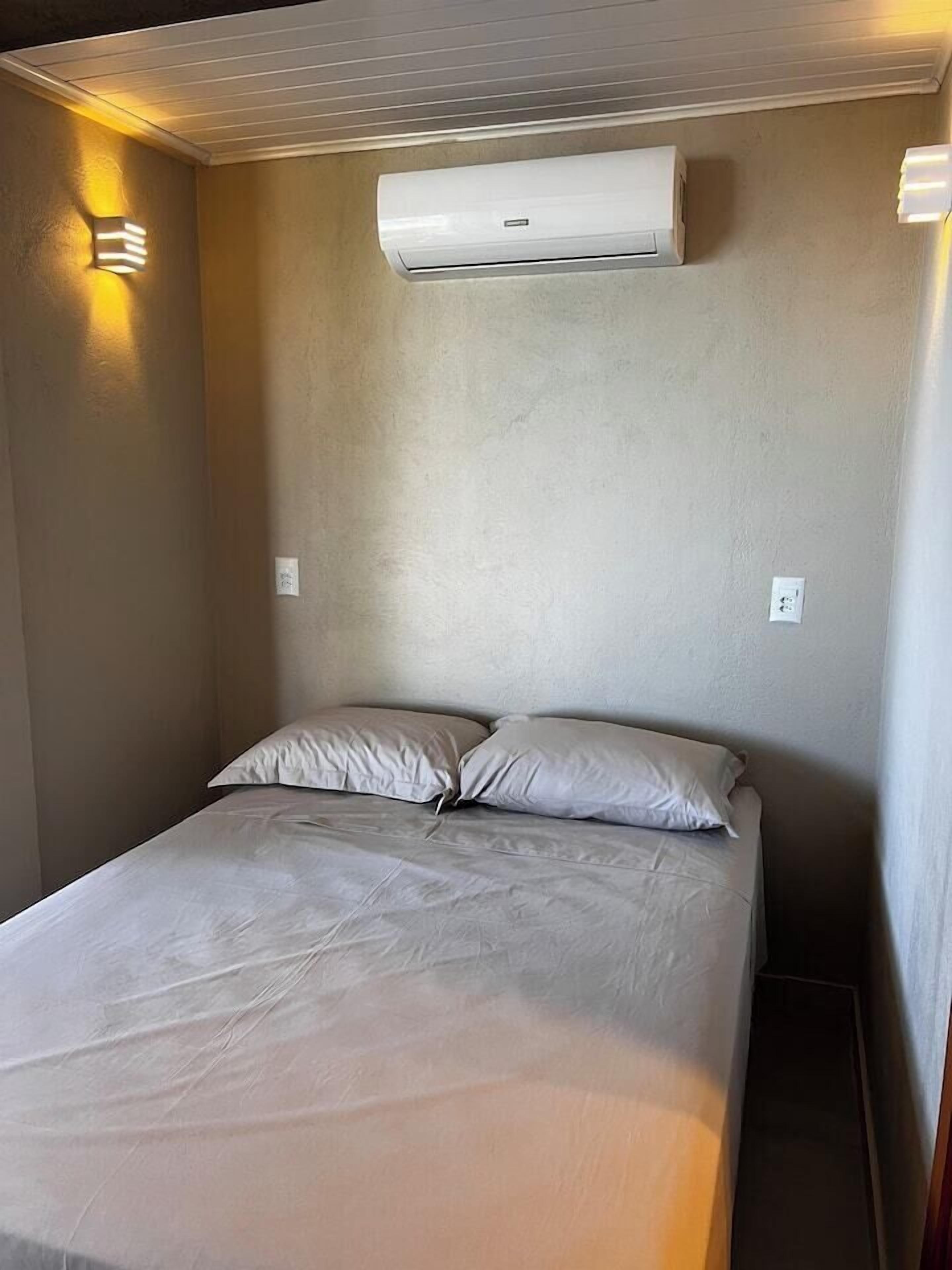 Piccola camera con un letto singolo, parete spoglia, climatizzatore a parete sopra il letto e lampada a muro a sinistra.