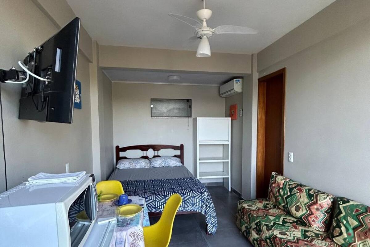 A compact hotel room with a bed, a desk, a chair, a sofa, a ceiling fan, and a TV mounted on the wall.