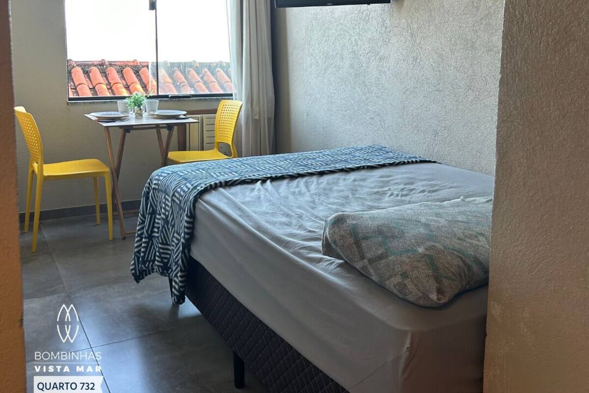 A bedroom with a bed, a small table with chairs, a window with curtains, and a wall-mounted TV.