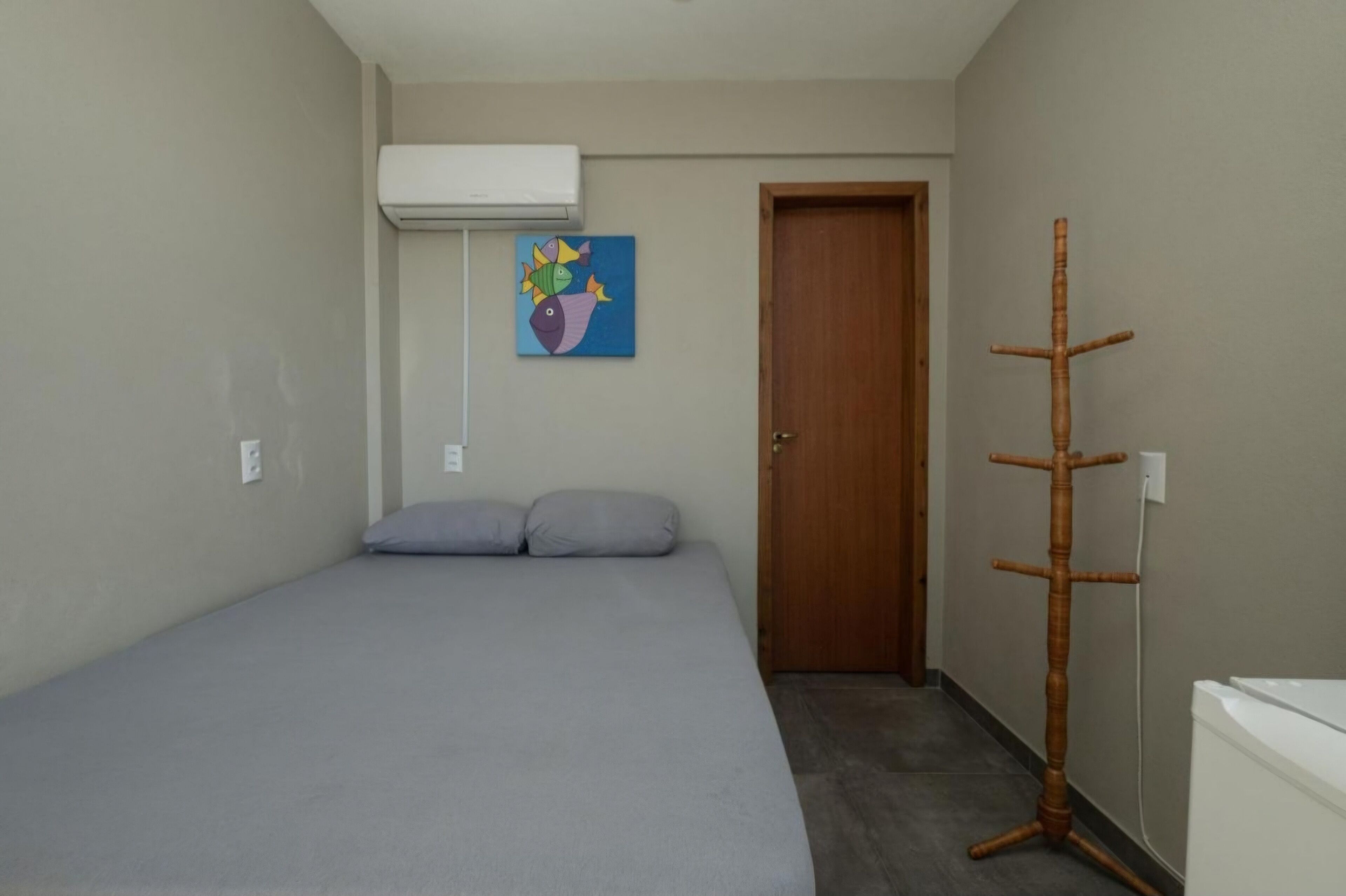 A small, neatly arranged bedroom with a bed, a wooden coat rack, a door, and a wall-mounted air conditioner.