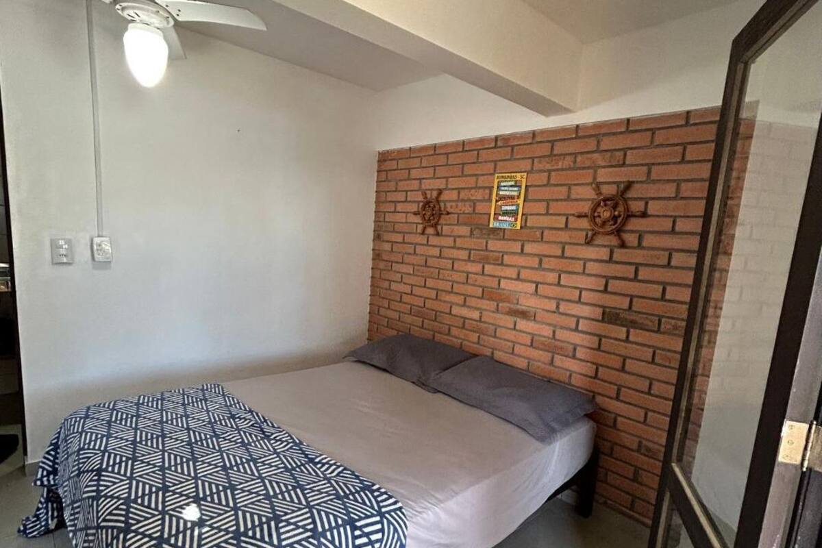 A bedroom with a brick accent wall, a bed with a patterned bedspread, a ceiling fan, and a window with blinds.
