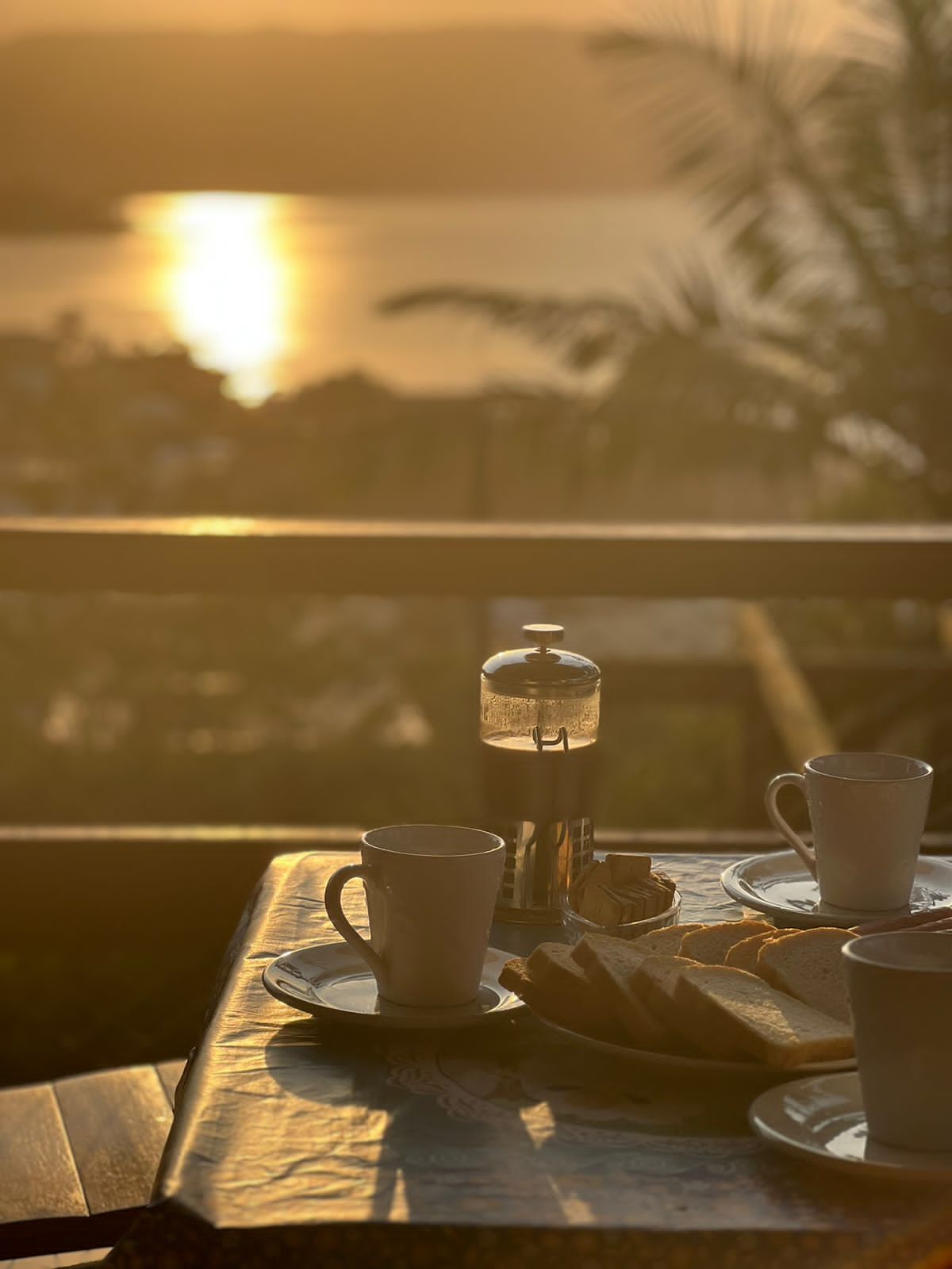 Un tavolo con una caffettiera francese, due tazze e un piatto con pane, sullo sfondo di una vista al tramonto.