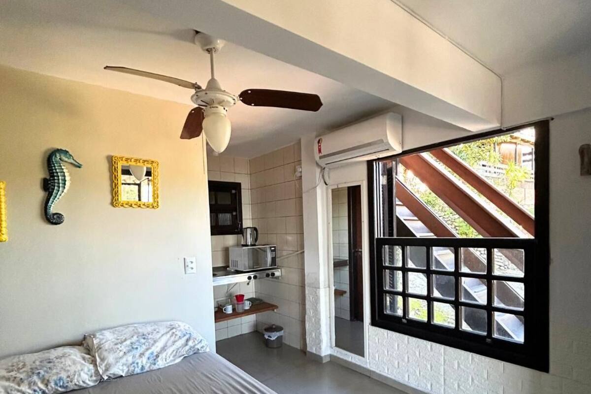 A bedroom with a bed, a ceiling fan, a window with a view of the outside, and a kitchenette area.