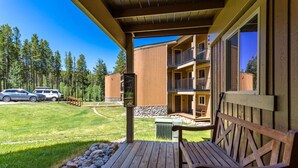 A wooden deck with a bench, a building with balconies, and a clear view of a forested area.