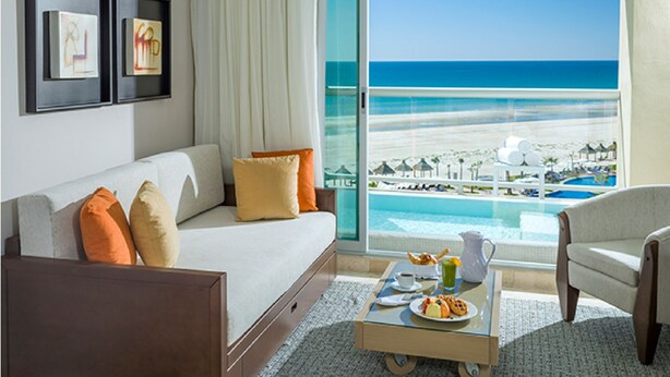 A hotel room with a large window overlooking a beach and pool, a sofa with cushions, a coffee table with breakfast items, and framed artwork on the wall.