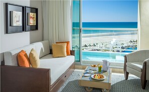 Camera d'albergo con un'ampia finestra vista spiaggia e piscina, un divano con cuscini, un tavolino con prodotti per la colazione e quadri appesi al muro.