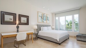 Une chambre moderne avec un lit, un bureau et des chaises, de grandes fenêtres offrant une vue dégagée et des œuvres d’art accrochées aux murs.