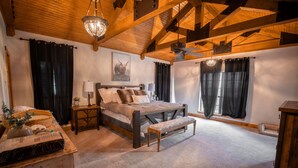 Un dormitorio con techo de madera, una cama con un banco, una mesita de noche, una lámpara y una ventana con cortinas.
