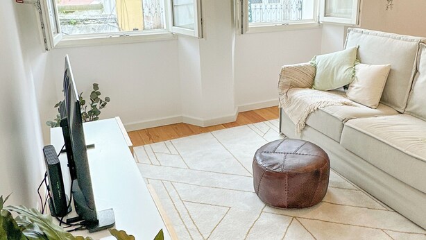 A living room with a sofa, ottoman, and a TV stand. There are two open windows with curtains, and a balcony is visible through one window. - Bombarda 15 Evora (Évora)