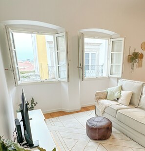 A living room with a sofa, ottoman, and a TV stand. There are two open windows with curtains, and a balcony is visible through one window. - Bombarda 15 Evora (Évora)