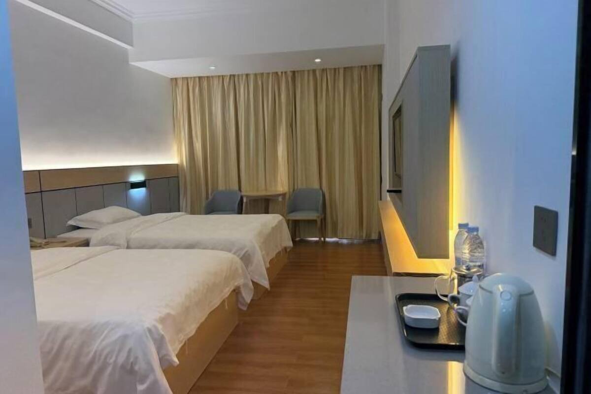Quarto de hotel com duas camas, uma mesa com chaleira, uma cadeira e uma janela grande com cortinas.