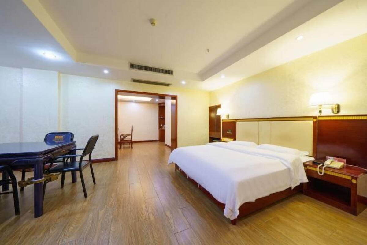 Quarto de hotel com piso de madeira, cama com roupa de cama branca, escrivaninha com cadeira e mesa de cabeceira com abajur e telefone.