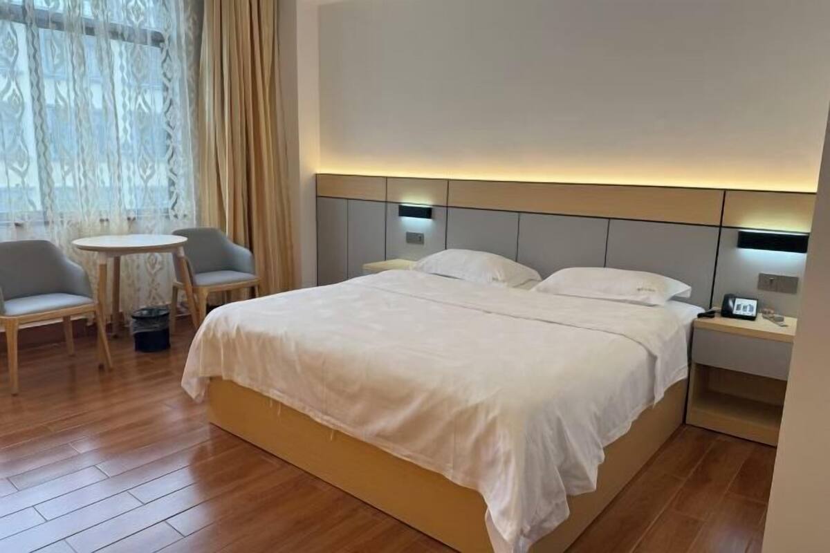 Quarto de hotel com uma cama grande, mesas de cabeceira, uma pequena mesa redonda com cadeiras e uma janela com cortinas.