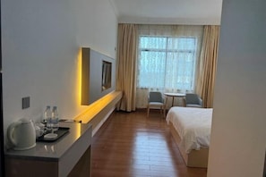 Habitación de hotel con cama, escritorio con tetera y botellas de agua, una silla y ventana con cortinas.