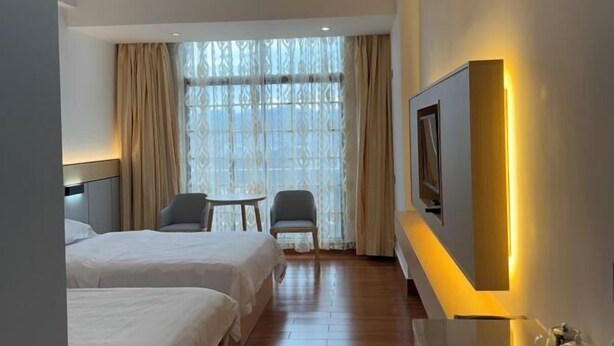 Habitación de hotel con dos camas, un escritorio, una silla, un ventanal grande con cortinas y un televisor de pared.