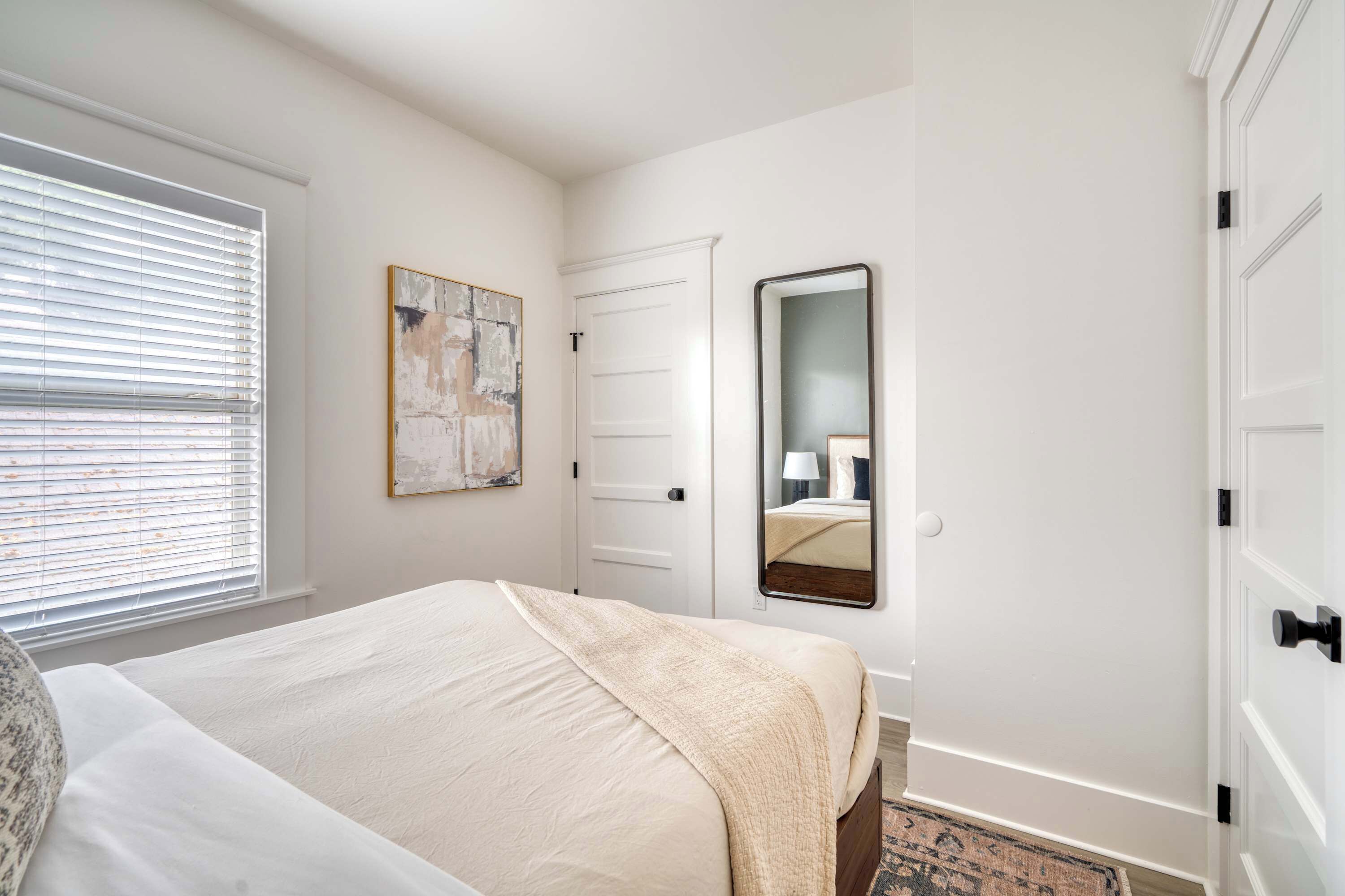 A bedroom with a bed, a window with blinds, a door, a mirror, and a painting on the wall.