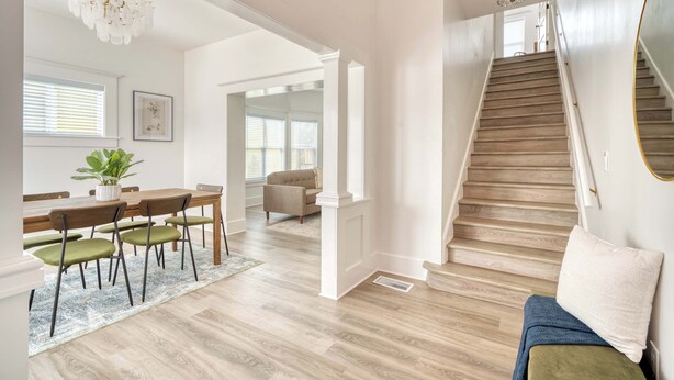 A spacious room with a wooden staircase, a dining area with a table and chairs, and a large window with blinds. - Victorian Vibes • 3BR • Steps to Mississippi (Portland)