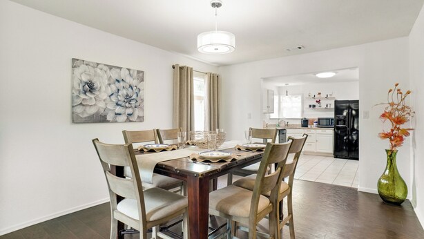 Een eethoek met een houten tafel gedekt voor zes personen, omringd door zes stoelen met beklede zitplaatsen. Aan de muur hangt een groot abstract bloemschilderij en op de vloer staat een groene vaas met bloemen.